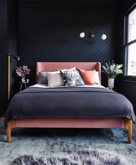 a small modern bedroom with black wooden walls, a pink bed and grey and pink bedding and greenery and blooms