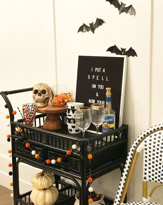 a Halloween bar cart styled with a pompom garland, a skull, a sign, pumpkins and some bats over it
