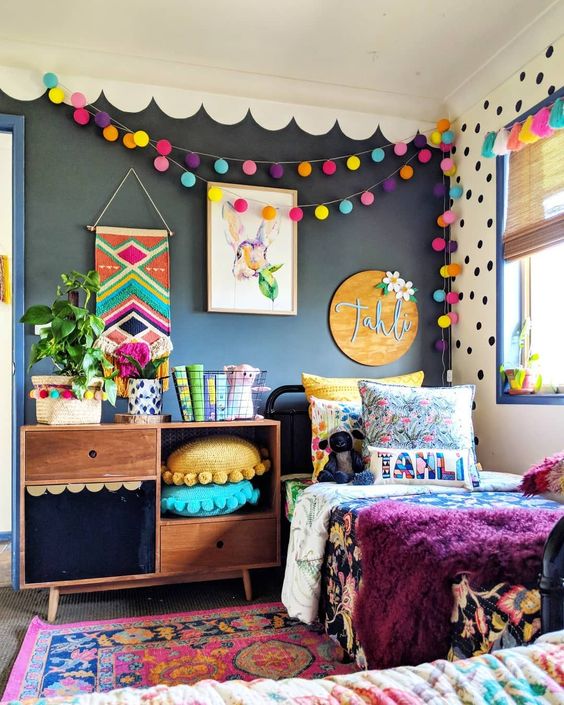 a bright kid's room with a black wall, colorful pompom garlands, pillows, bedding and a rug is extra bold