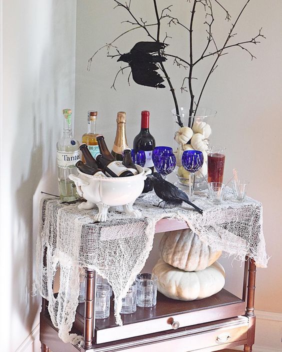 a colorful Halloween bar cart with cheesecloth, neutral pumpkins, blue glasses, a pumpkin vase with branches and a crow