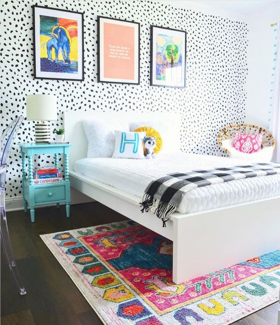 a colorful kid room with a Dolmatin wall, colorful bedding and a rug, bold artworks, a blue nightstand and a rattan chair