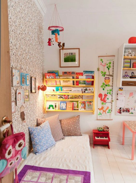 a colorful kid's room with a wallpaper wall, a yellow shelf, bright artworks and bedding and colorful toys
