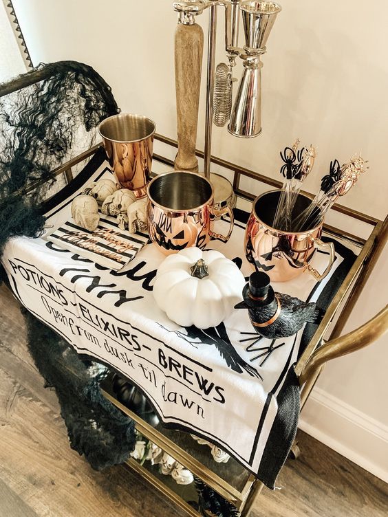 a cool Halloween bar cart with copper mugs, a crow, white pumpkins, skulls, black spiderwebs and shiny touches
