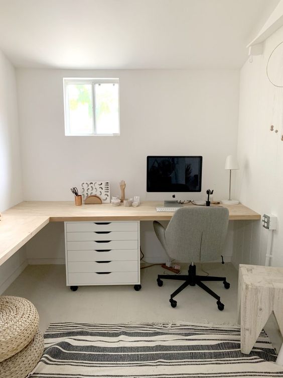 a neutral farmhouse home office with an L-shaped desk, drawers, a grey chair, a stool, jute poufs, some decor and a lamp