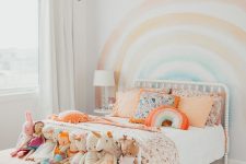 a pastel rainbow kid’s room with a rainbow on the wall, colorful bedding and peachy stools with toys
