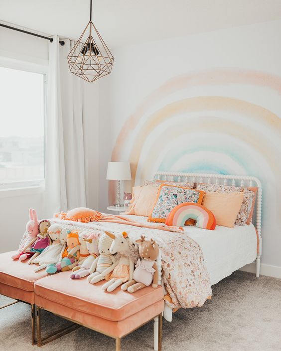 a pastel rainbow kid's room with a rainbow on the wall, colorful bedding and peachy stools with toys