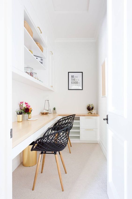 a small white home office with an L-shaped desk with storage, black chairs, a storage unt, some decor and blooms