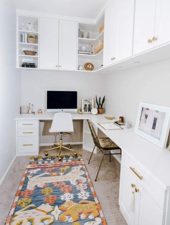 a white boho home office with an L-shaped desk with storage, kitchen cabinets for storage, a bold rug and mismatching chairs