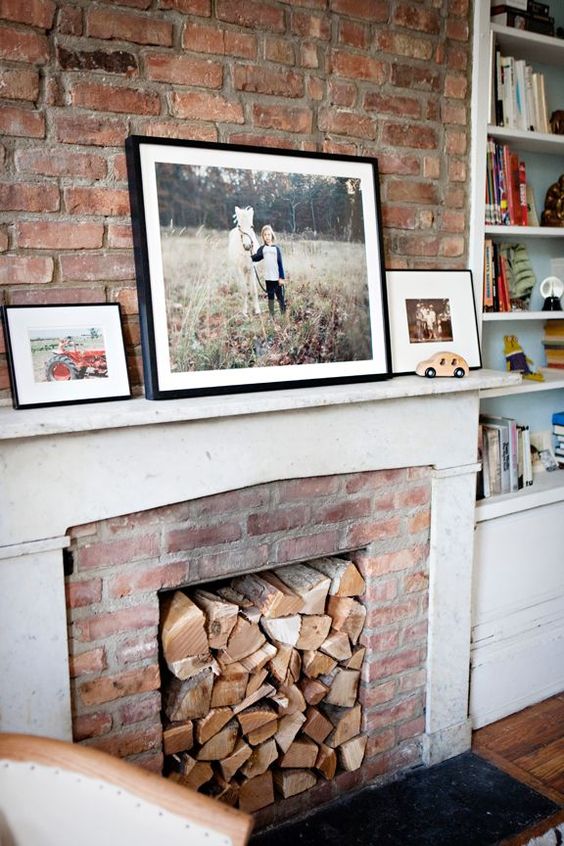 a rustic fireplace of red brick, with a white mantel and some firewood inside the fireplace plus photos on the mantel