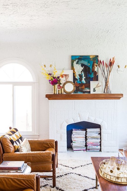 a beautiful white living room with amber leather chairs, a vintage white fireplace used for book storage, with colorful artworks and blooms