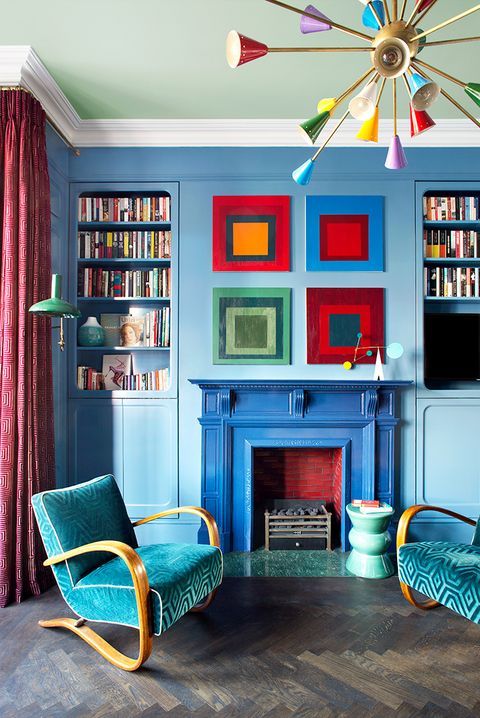 a blue living room with built-in bookcases, a bright fireplace, teal printed chairs, bold artwork and a colorful chandelier