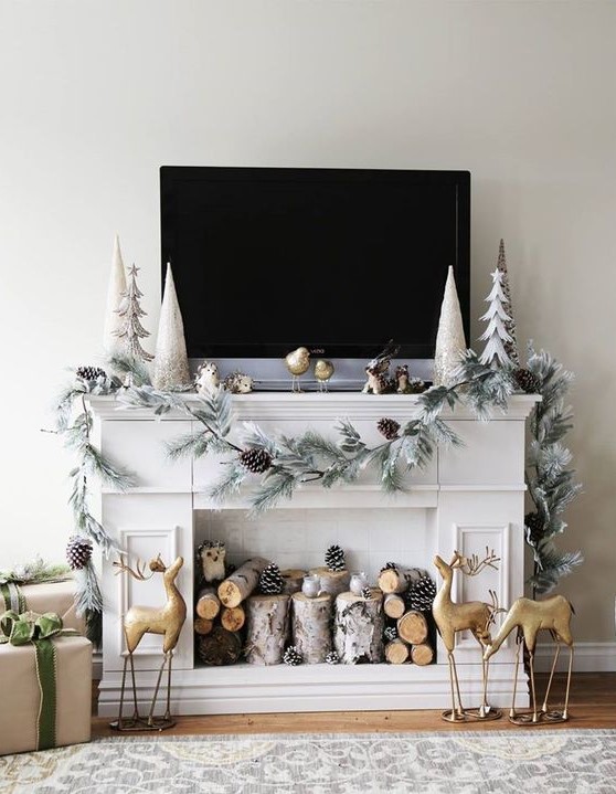 a faux fireplace styled for Christmas, with firewood, pinecones, gold deer, a whitewashed evergreen garland with pinecones and pretty trees