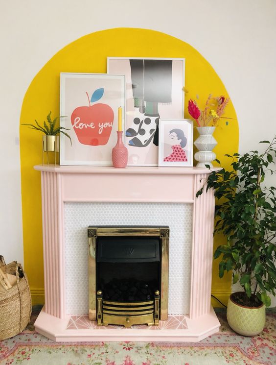 a fireplace with a blush mantel and a bold yellow accent on the wall plus bright decor on the mantel is cool