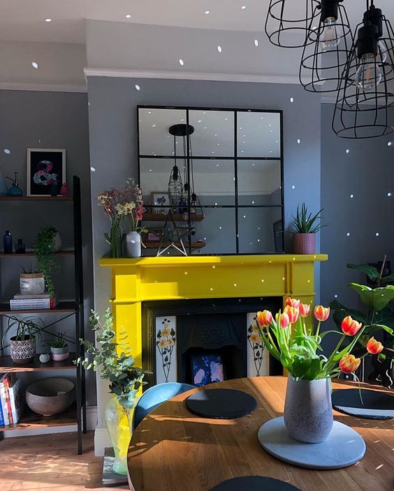 a grey living room with a fireplace clad with a yellow mantel, with pendant lamps, a table with some porcelain and some blooms