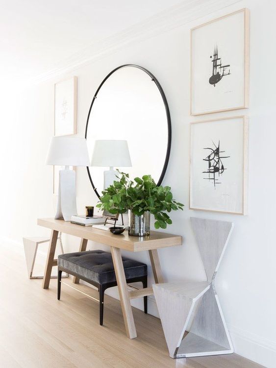 a modern entry with a round mirror, a wooden console, an upholstered ottoman, matching geometric chairs and greenery