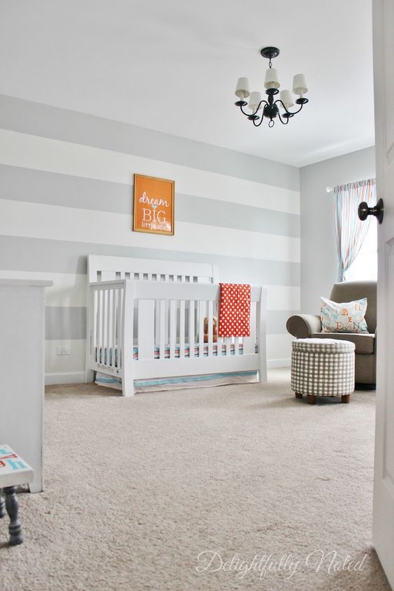 a neutral and very simple nursery with a grey and white striped wall, a retro chandelier, a crib and an ottoman
