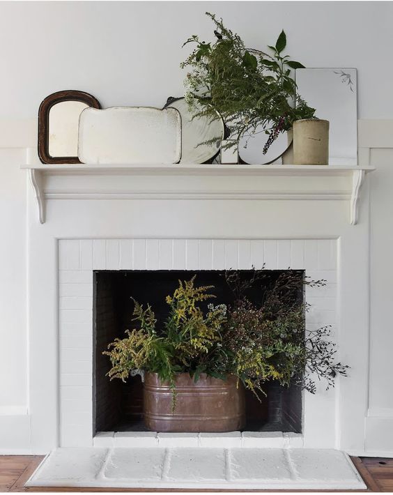 a non-working fireplace with a bucket with evergreens, an arrangement of mirrors and greenery on the mantel