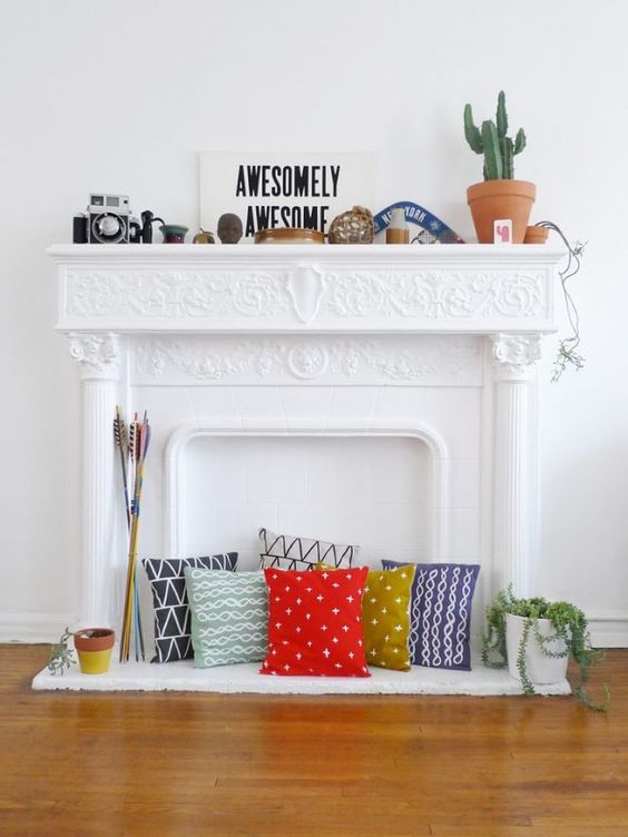 a non-working fireplace with an ornated mantel, with colorful pillows, some vintage decor and potted plants