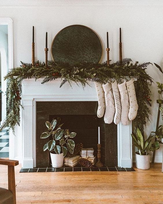 a non-working fireplace with books, pinecones, a potted plant and a candle, evergreens, candles and stockings