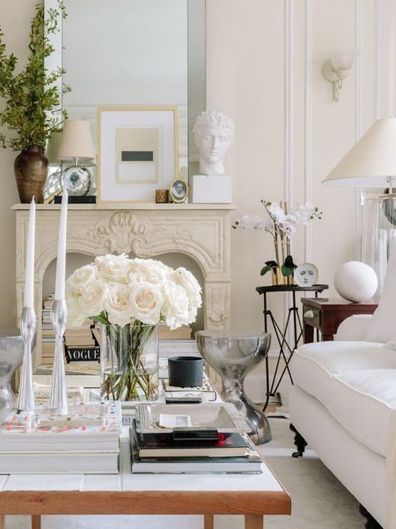 a refined neutral living room with creamy walls and furniture, with a low coffee table, a chic mantel and books in the fireplace plus an oversized mirror