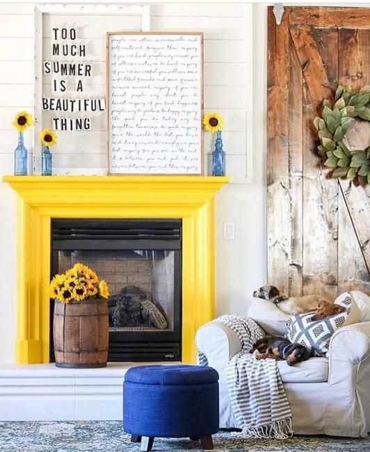 a rustic living room with a fireplace and a yellow mantel, a neutral chair, a navy pouf, some rustic decor