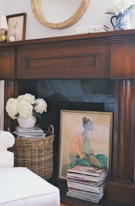 a stylish fireplace with a dark stained mantel, a stack of magazines, an artwork and a basket with books and blooms