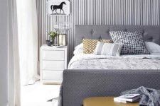 a stylish grey and white bedroom with a striped accent wall, a grey bed, a mustard ottoman and artworks