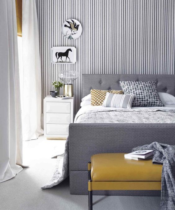 a stylish grey and white bedroom with a striped accent wall, a grey bed, a mustard ottoman and artworks