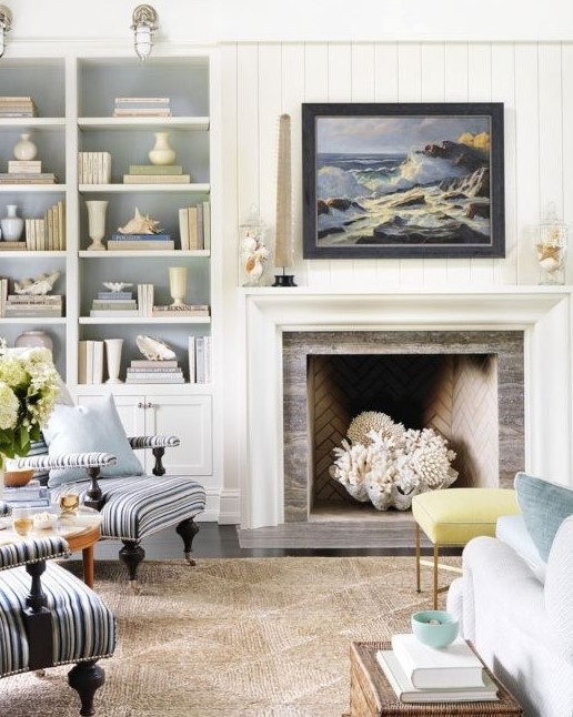 a white coastal living room with built-in shelves and a storage unit, a fireplace with a giant seashell and corals, light blue and striped furniture