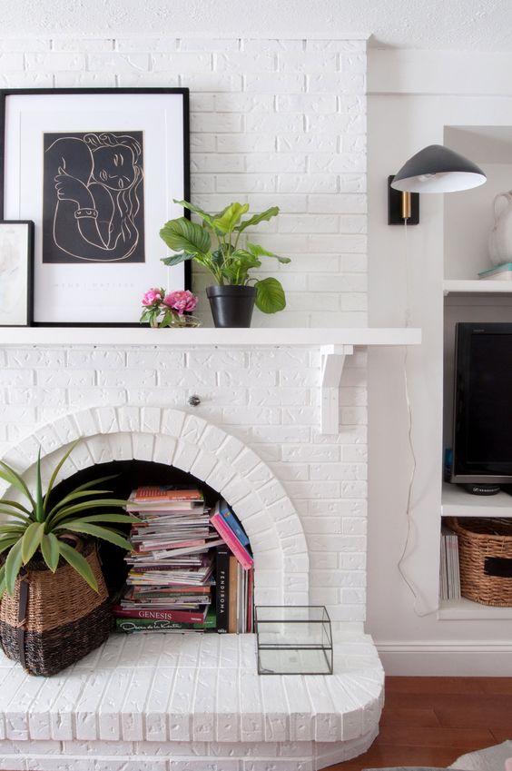 an arched fireplace with books and magazines and a potted plant, an artwork and some blooms on the mantel