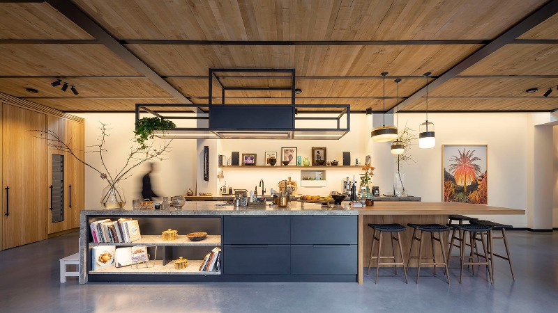 The kitchen is done with dark metal cabinets, there's a small wooden breakfast bar, with many lights and wooden stools