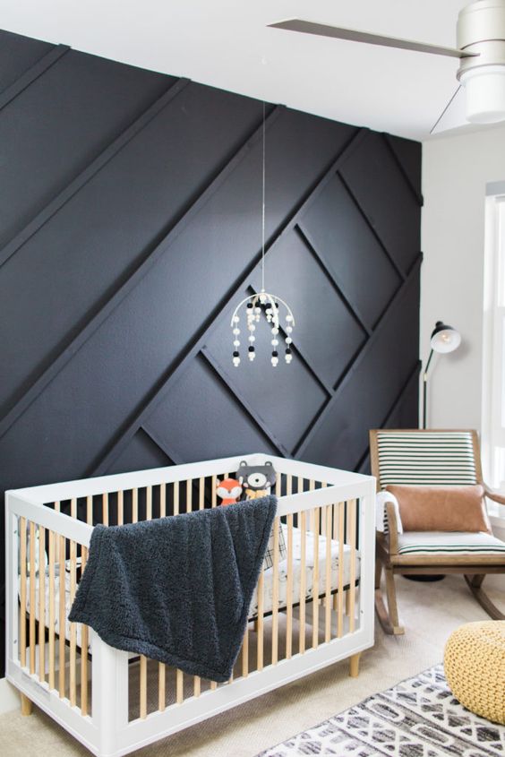 a bold nursery with a black geometric paneled wall, light-colored wooden furniture, geometric bedding and a cute mobile