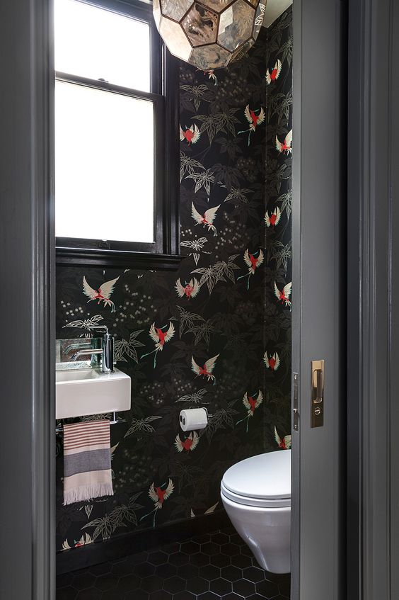 a moody and refined restroom with black fauna print wallpaper, blakc tiles on the floor and a refined gold faceted lamp