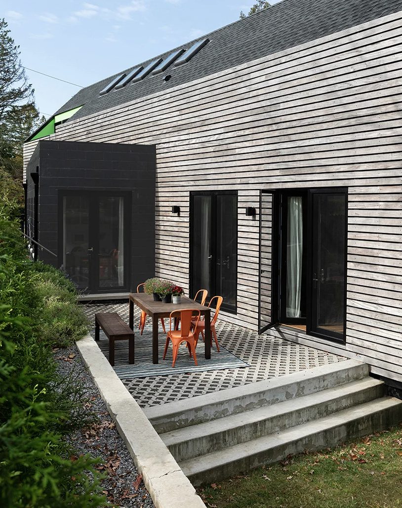 The exterior of the house is contrasting, dark and light, with a terrace with a wooden table and orange chairs