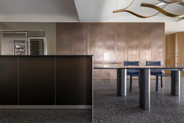 The kitchen is done with sleek dark and metallic cabinetry, there's a large table and a couple of blue chairs The kitchen is done with sleek dark and metallic cabinetry, there's a large table and a couple of blue chairs