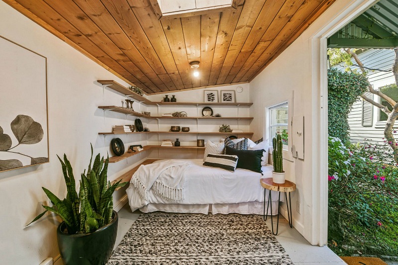 The guest cottage features a modern boho bedroom with floating shelves, some potted plants and a wooden ceiling