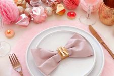 a pink holiday tablescape with pink chargers, ornaments, paper pompoms, glasses, candles and napkins plus matte cutlery