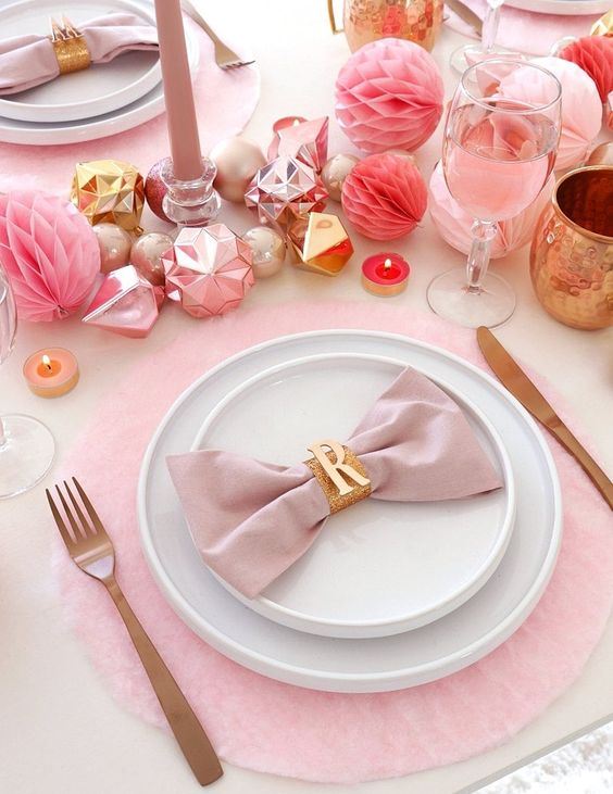 a pink holiday tablescape with pink chargers, ornaments, paper pompoms, glasses, candles and napkins plus matte cutlery