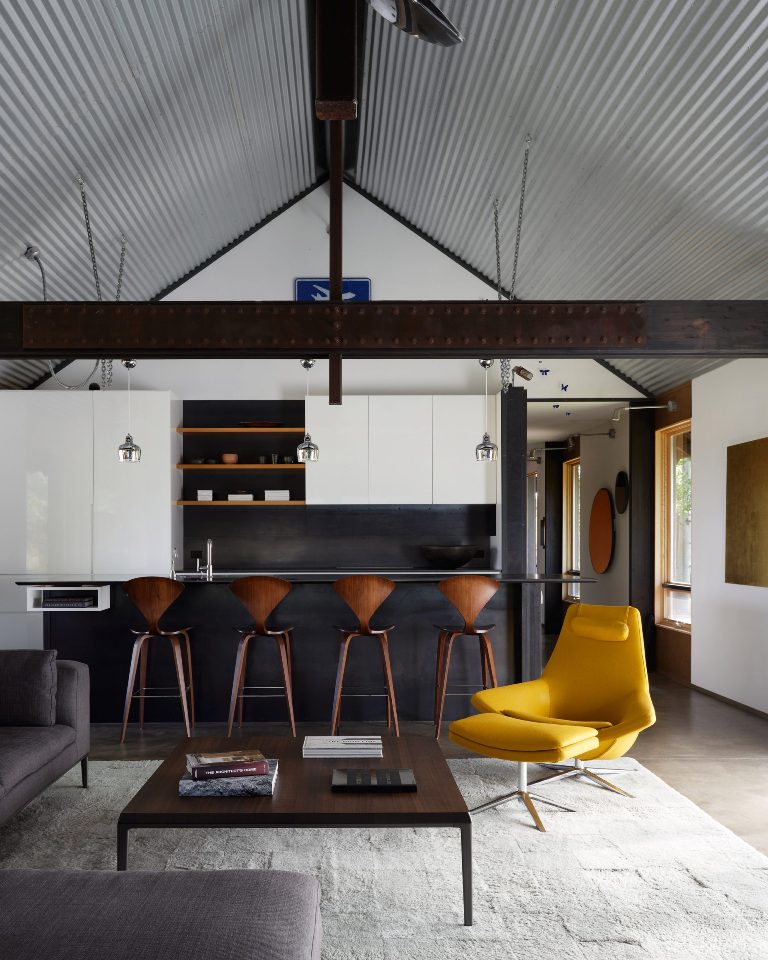 The open layout features a kitchen, a dining space and a living room, with sleek white cabinets, a grey sectional and a yellow lounger