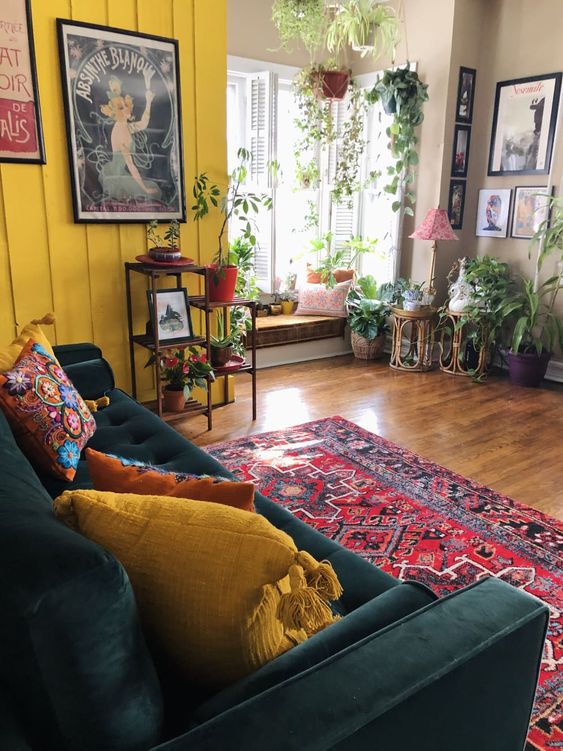 a boho living room with a mustard wall, a dark green sofa, bright and printed pillows, potted greenery, statement artworks