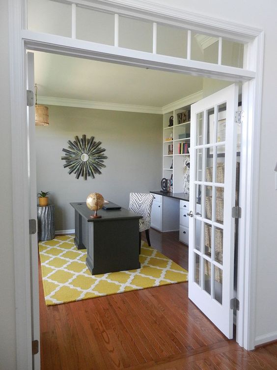 a bold home office with a grey accent wall, a white storage unit that takes a whole wall, a black desk, a bright printed rug and a sunburst mirror