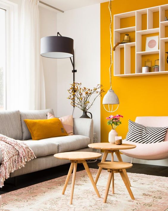 a chic contemporary living room with a marigold accent wall and a pillow, neutral and pastel furniture, a floor lamp and mini wooden tables