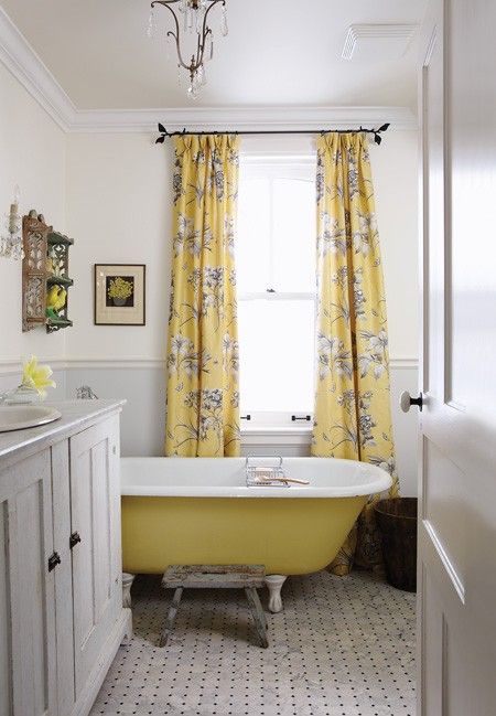 a chic vintage bathroom with grey paneling, a yellow bathtub, yellow floral curtains, printed tiles and a crystal chandelier