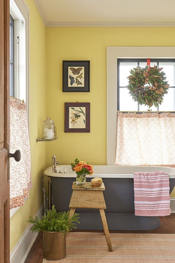 a farmhouse bathroom with yellow walls, a grye bathtub, printed textiles, some artworks and greenery and foliage