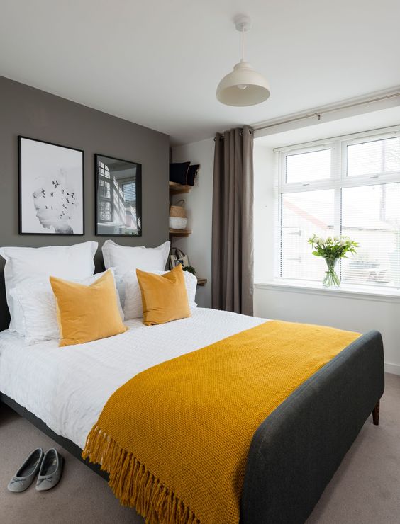 a grey and white bedroom with a grey upholstered bed, white and mustard bedding, a pendant lamp and a gallery wall that adds chic to the space