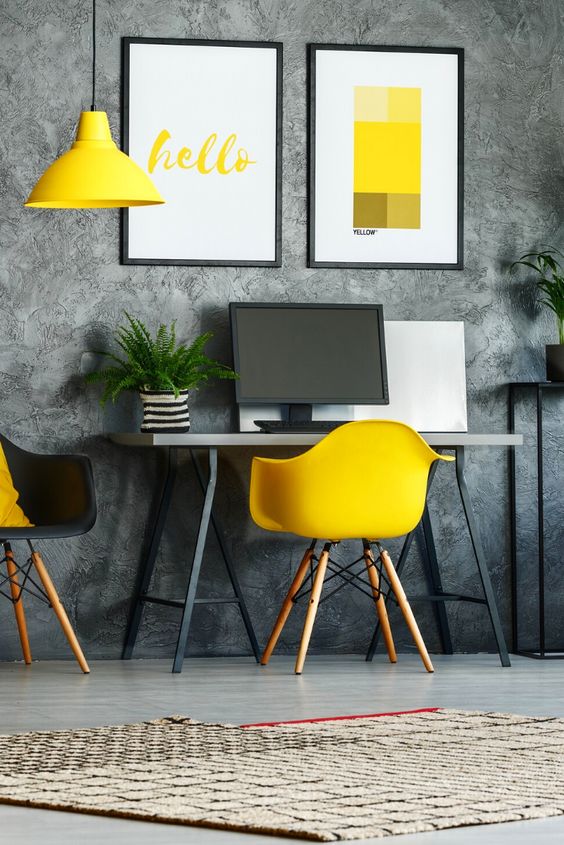 a modern home office with a grey wallpaper wall, a small desk, a black and a yellow chair, a yellow pendant lamp and some bold prints