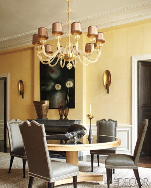 a refined and chic grey and yellow dining room with yellow paneled walls, a round table, grye chairs, a chic chandelier and a marble fireplace
