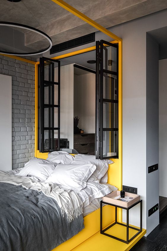 a stylish minimalist bedroom with dove grey walls, a window to a bathroom, a sleeping zone accented with bold yellow