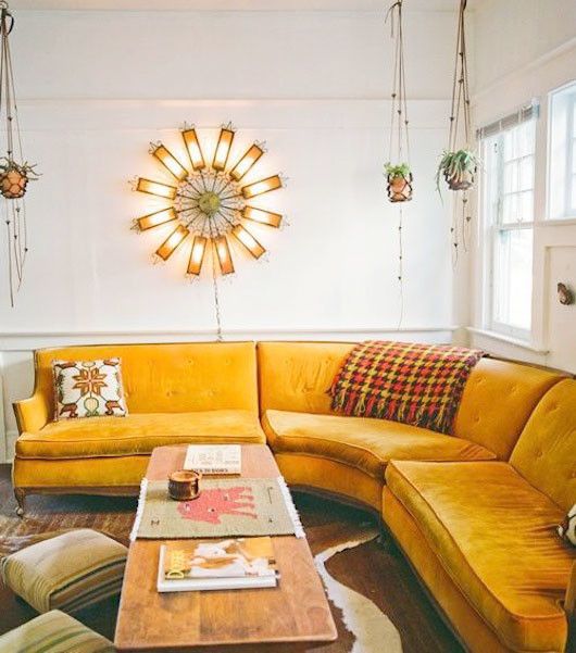 a welcoming boho living room with a curved honey yellow sofa, a wooden table, striped stools, a sunburst lamp and hanging plants