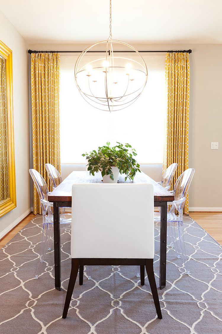 an eclectic and chic dining rom with a wooden table, acrylic and upholstered chairs, a sphere pendant lamp, yellow curtains and a large mirror